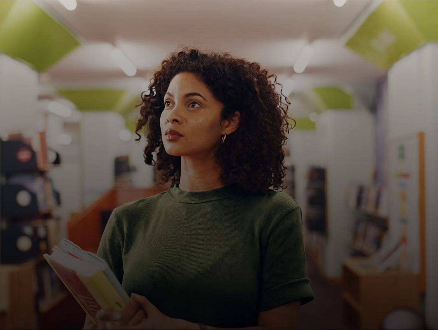 Young woman with curly hair holding books in a library, looking thoughtfully to the side.