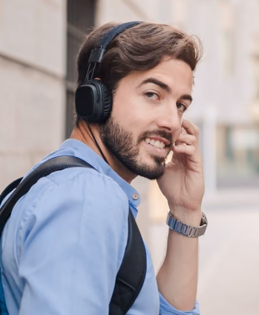 Young man with beard wearing headphones and backpack smiling while walking outdoors.