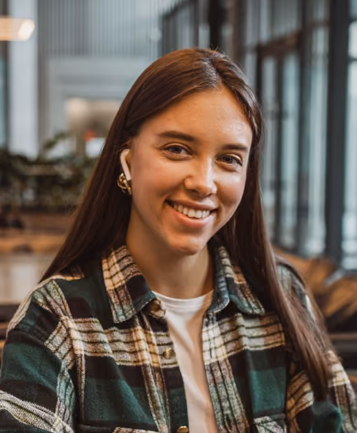 Smiling young woman with long brown hair wearing AirPods and a plaid shirt indoors.