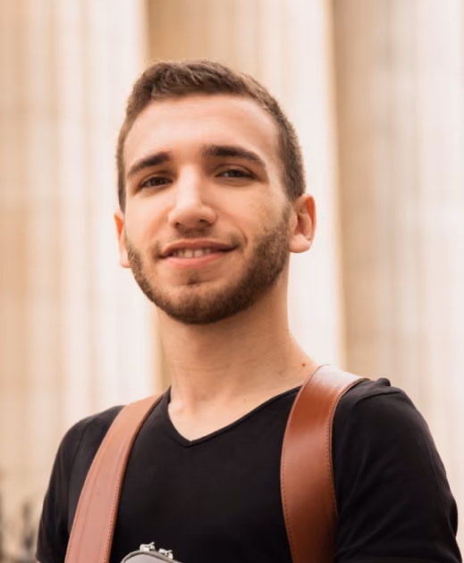 Young man with short hair and beard wearing a black shirt and brown straps, smiling outdoors.