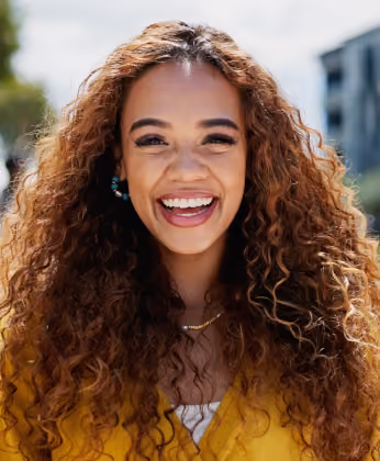 Smiling woman with long curly hair wearing a yellow top outdoors on a sunny day.
