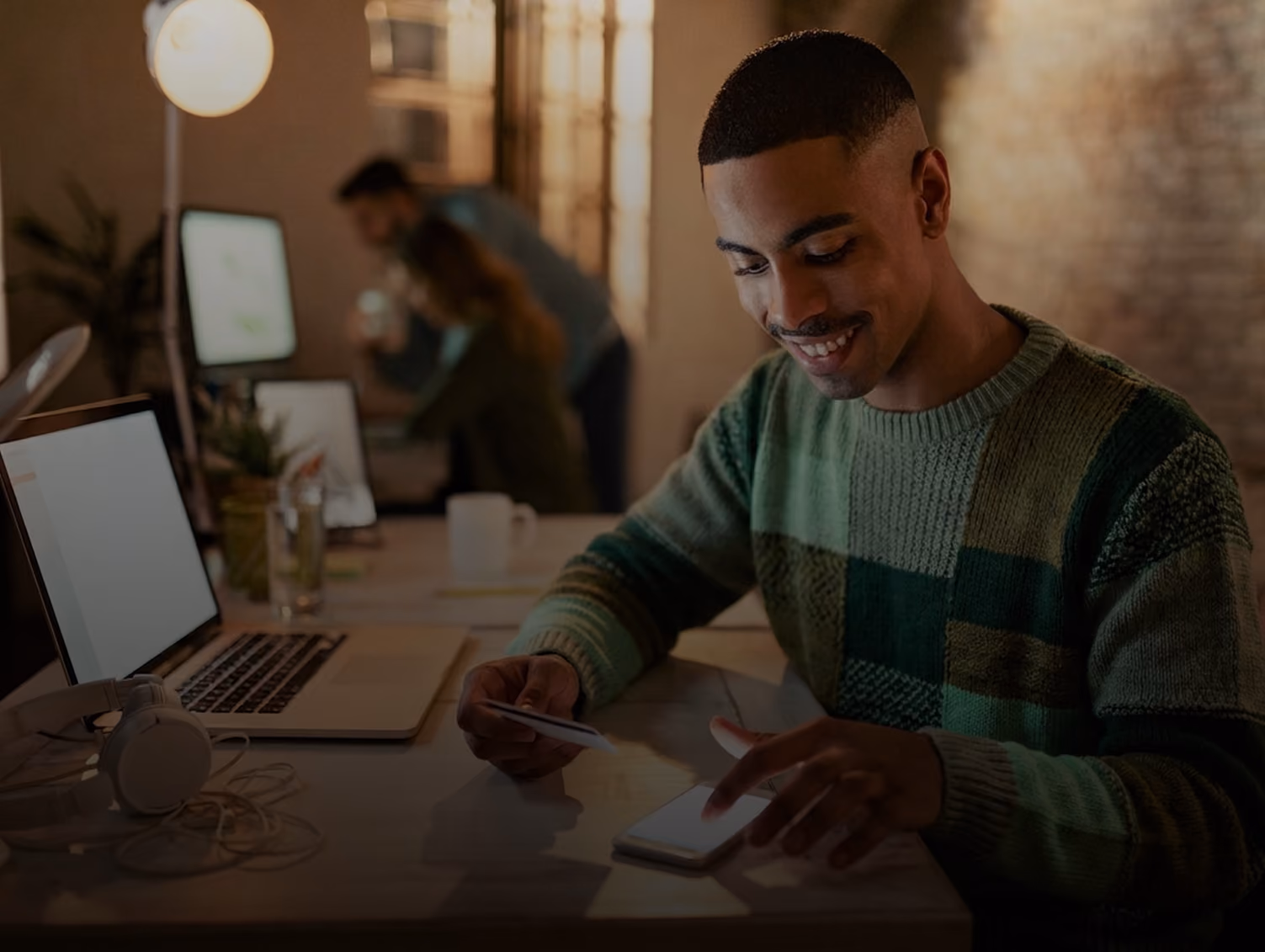 Man smiling while using smartphone and holding credit card at desk with laptop and headphones.