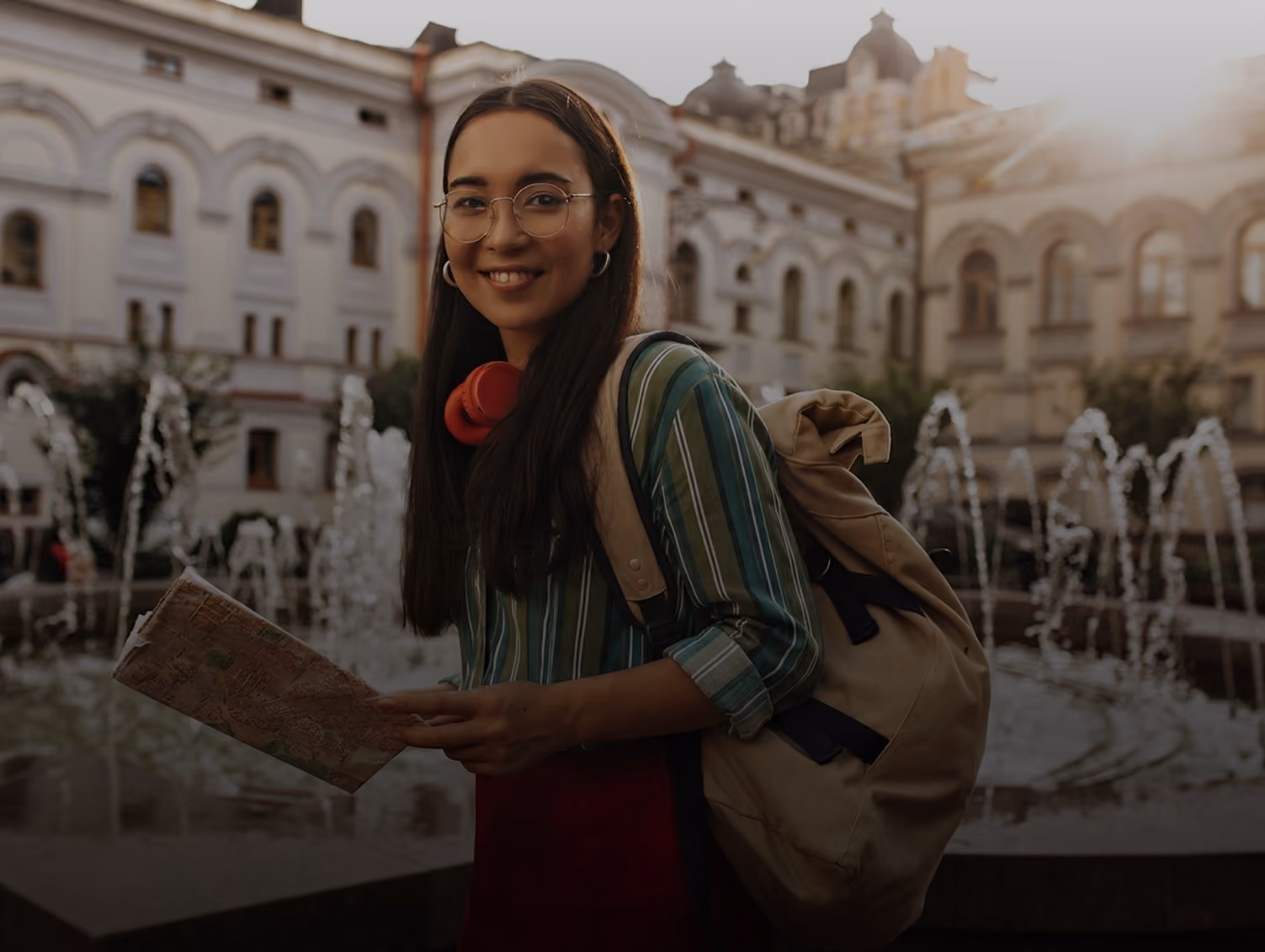 Smiling woman with glasses, backpack, and map stands near fountains in a historic city square at sunset.